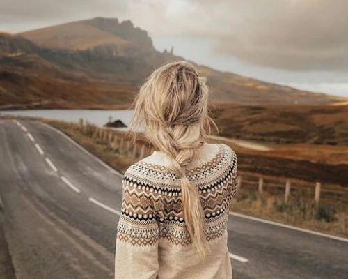 A person with braided hair stands on a road, overlooking a scenic landscape with hills and a lake in the background.