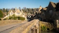 Charming Cotswold village with stone cottages lining a quiet street, framed by lush greenery under a clear blue sky.