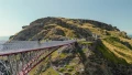 A scenic view of a pedestrian bridge connecting cliffs, with people walking and a coastline in the background under a clear sky.
