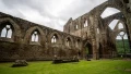 Ruins of an ancient stone abbey with Gothic arches, set against a cloudy sky and surrounded by green grass.