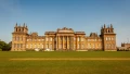 Blenheim Palace's grand facade stands against a clear blue sky, surrounded by lush green lawns, showcasing its stunning architecture.