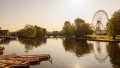 A serene riverside scene with boats, a ferris wheel, and lush trees reflecting on the water under a warm sunset glow.