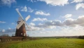 A rustic windmill stands tall in a lush green field under a bright blue sky with fluffy clouds.