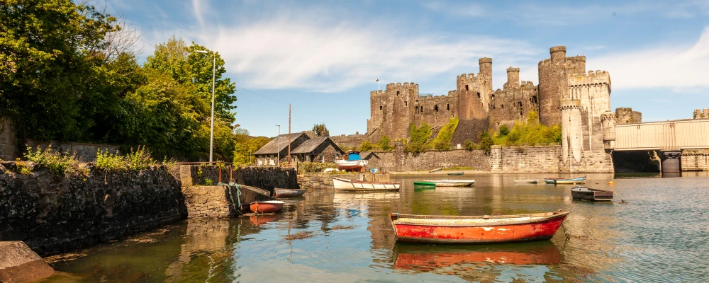 A scenic view of a castle by the water, surrounded by greenery and colorful boats reflecting in the calm water.