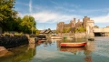 A scenic view of a castle by the water, surrounded by greenery and colorful boats reflecting in the calm water.