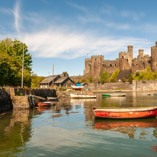 A scenic view of a castle by the water, surrounded by greenery and colorful boats reflecting in the calm water.