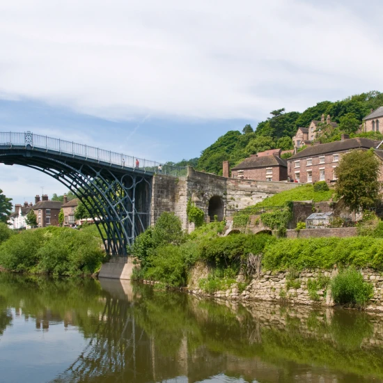 Iron bridge spanning a river, with lush greenery and historic buildings along the banks under a clear blue sky.