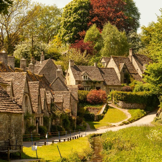 Quaint stone cottages nestled among lush greenery and colorful trees, with a winding path and stream in a tranquil rural setting.