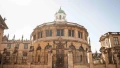 A historic architectural building with a green dome, ornate details, and surrounding sculptures in bright sunlight.