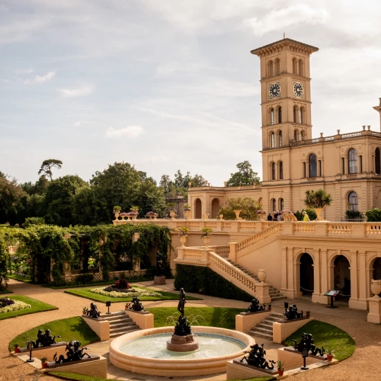 A grand building with a clock tower overlooks beautifully landscaped gardens featuring sculptures and fountains.