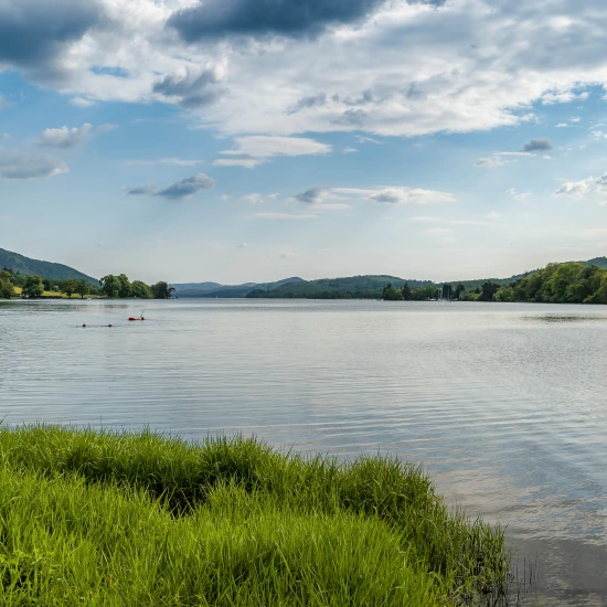 Scenic view of a calm lake surrounded by greenery and rolling hills under a cloudy sky, ideal for nature tours.