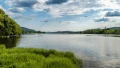 Scenic view of a calm lake surrounded by greenery and rolling hills under a cloudy sky, ideal for nature tours.
