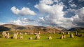 A panoramic view of standing stones in a green field, surrounded by rolling hills and a dramatic sky with clouds.
