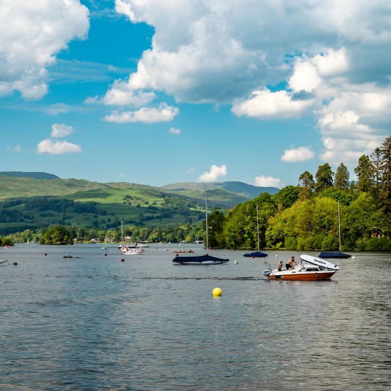 Scenic view of a lake with boats, surrounded by lush greenery and hills under a bright, partly cloudy sky.