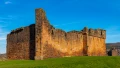Historic stone castle ruins against a clear blue sky, surrounded by green grass with residential buildings in the background.