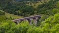 A scenic view of a historic stone viaduct surrounded by lush green hills and trees, with cyclists traversing the bridge.