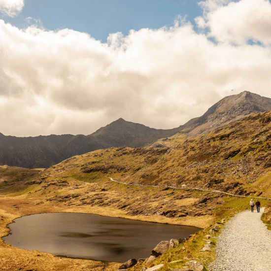 A scenic pathway winds through rugged mountains beside a tranquil lake under a partly cloudy sky. Ideal for small group tours.