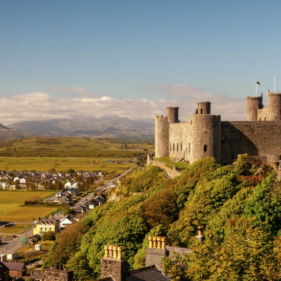 A scenic view of a stone castle surrounded by greenery, hills, and a village, under a clear blue sky in the UK.