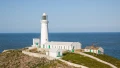 A white lighthouse stands on a cliff overlooking the blue ocean, surrounded by green grass and coastal buildings.