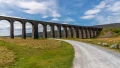 A scenic view of a stone viaduct towering over a grassy landscape under a blue sky, with a winding gravel path nearby.