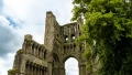 Historic stone ruins with large arches, surrounded by lush greenery under a cloudy sky.