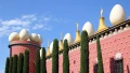 Colorful building topped with large eggs and golden statues, surrounded by tall cypress trees against a blue sky.