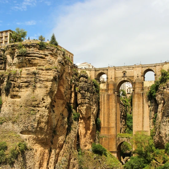 A stunning view of a bridge spanning between high cliffs, framed by lush greenery and quaint buildings above.