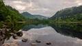 A serene lake reflecting lush green hills and a cloudy sky, framed by rocks along the water's edge.