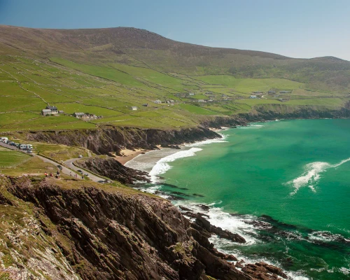 Scenic view of lush green cliffs meeting a turquoise sea, with scattered houses and a winding road along the coastline.