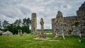 Ancient stone structures and crosses in a grassy field, surrounded by trees under a cloudy sky.