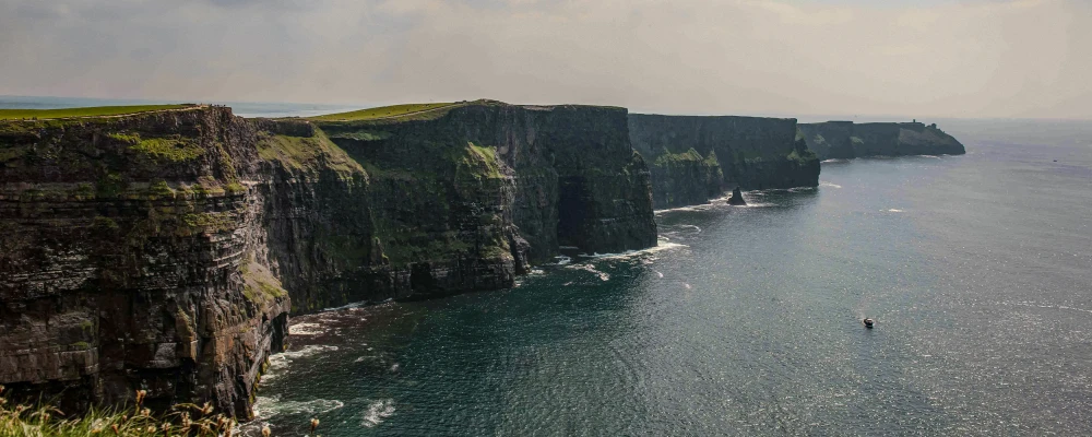 Scenic view of rugged cliffs meeting the ocean, with a small boat visible in the water under a partly cloudy sky.