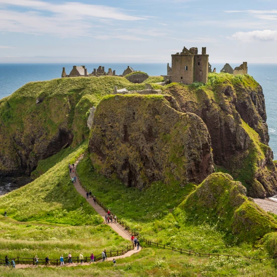 A scenic view of a coastal cliff with a historic ruin, surrounded by lush greenery and a pathway leading to the beach.