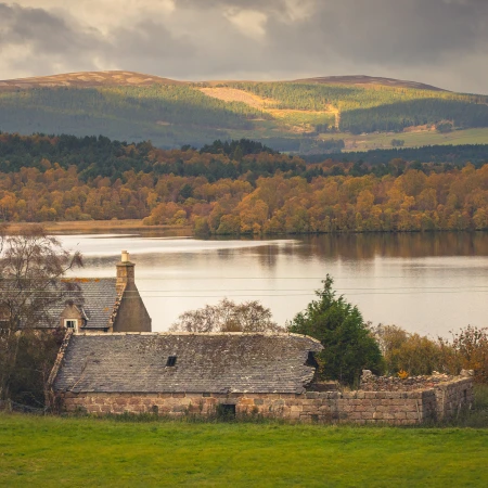 Picturesque landscape featuring a serene lake, lush autumn trees, and rustic buildings under a moody sky. Perfect for exploring.