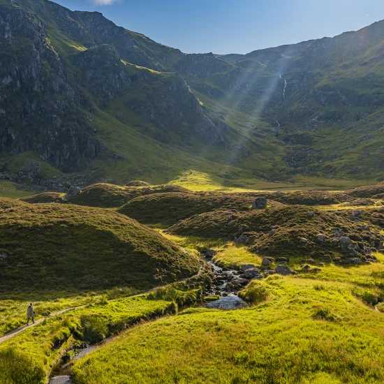 A scenic view of lush green hills and a stream, with a person walking along a path under a bright blue sky.