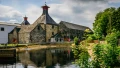 Historic stone buildings beside a tranquil river, surrounded by lush greenery and under a partly cloudy sky.