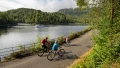 Three cyclists ride along a scenic path beside a calm lake, surrounded by lush greenery and distant hills.