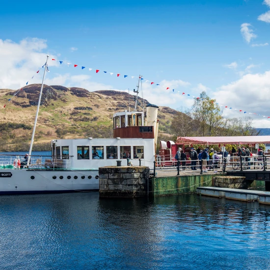 A scenic view of the boat "Sir Walter Scott" docked with passengers boarding, surrounded by hills and a clear blue sky.