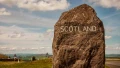 A large rock bearing the word "SCOTLAND" in white, set against a scenic landscape with clouds and gentle hills in the background.