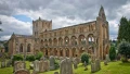 Ruins of an old stone abbey with arched windows, surrounded by a graveyard and greenery under a cloudy sky.