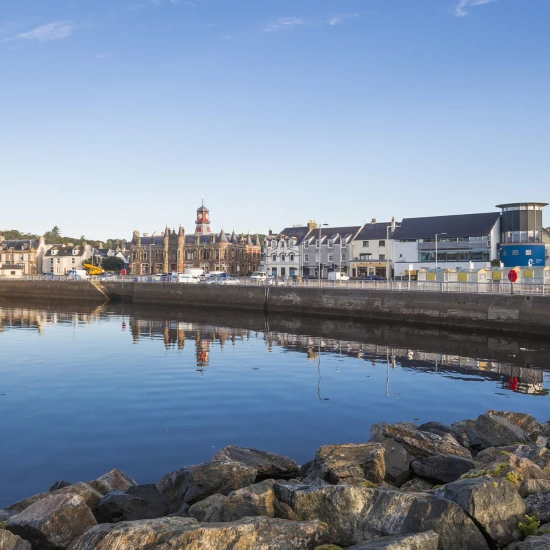 A tranquil waterfront scene showcasing buildings along the shore, reflecting in calm waters under a clear blue sky.