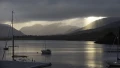 A serene view of a lake with boats, framed by dark mountains and clouds, illuminated by soft rays of sunlight breaking through.