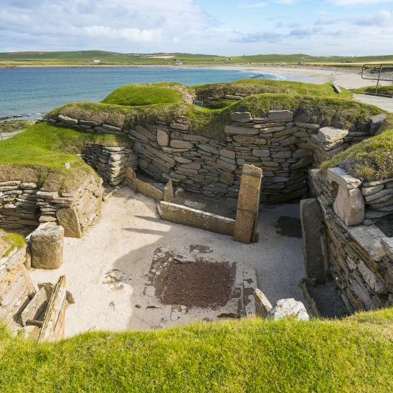 Ancient stone ruins surrounded by grassy mounds, overlooking a beach and ocean under a partly cloudy sky.
