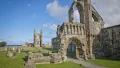 Ruins of an ancient stone abbey with towers and gravestones, set against a clear blue sky by the sea.
