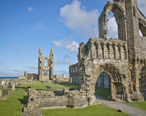 Ruins of an ancient stone abbey with towers and gravestones, set against a clear blue sky by the sea.