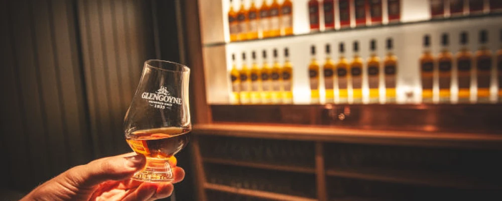 A hand holding a Glengoyne whisky glass in front of a stylish shelf filled with whisky bottles.