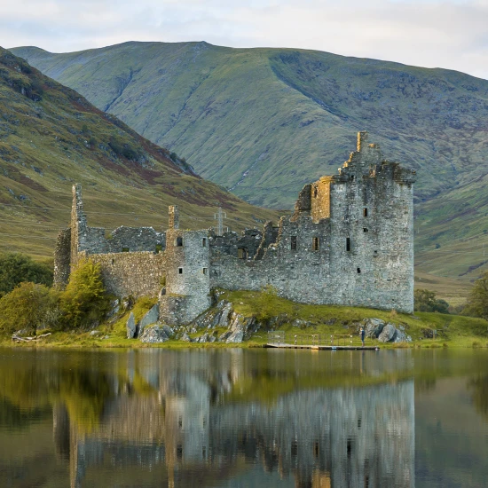 Ruins of a stone castle by a tranquil lake, surrounded by green hills under a cloudy sky. Reflective water enhances the scenery.