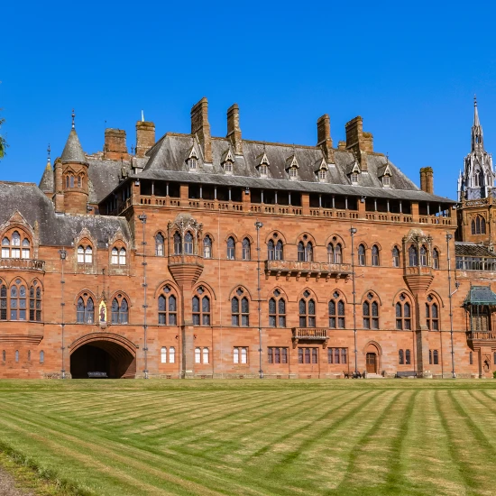 Historic red brick mansion surrounded by lush greenery and a clear blue sky, showcasing intricate architectural details.