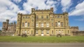 Historic stone castle with rounded turrets and garden in front, under a blue sky with clouds.