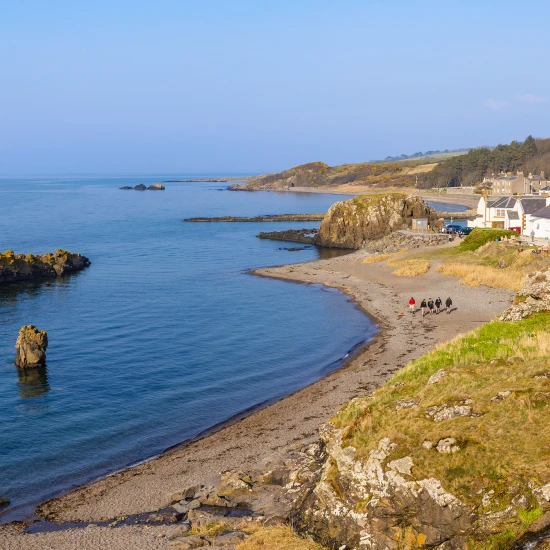 A serene coastal view featuring rocks, sandy beach, and a small group of people walking along the shoreline under a clear sky.