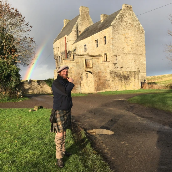 A man in a kilt stands near a historic castle, with a rainbow in the background and a scenic landscape around him.